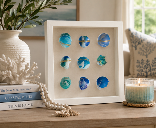 A framed decorative wall piece with nine blue and teal eggshell pieces arranged in a 3x3 grid on a beige background, displayed on a wooden table in a styled room with a vase, books, coral, bead garland, and a candle.