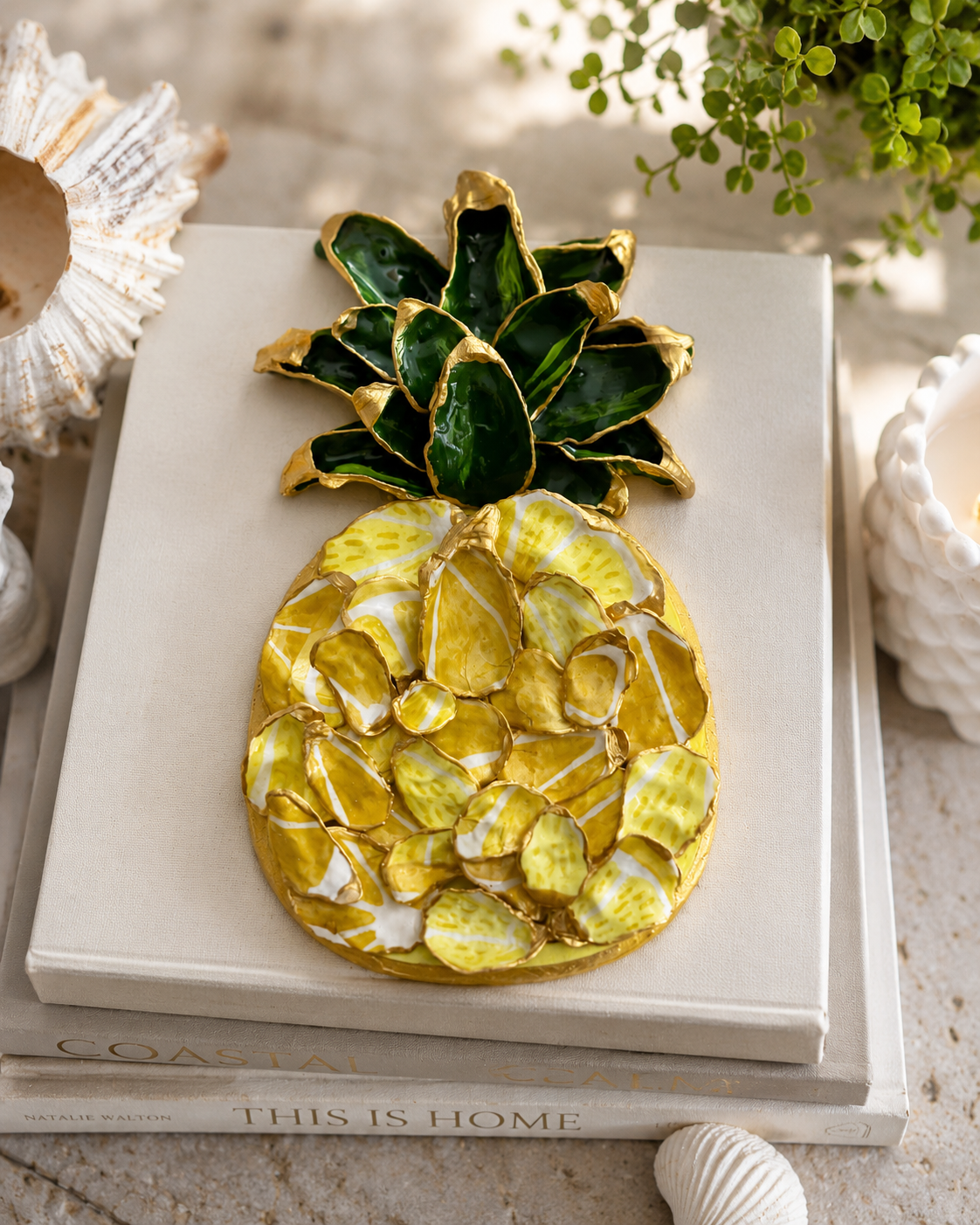 AI lifestyle image of The Golden Pineapple Shell Plaque displayed on a stack of books — coastal shell art with gold paint edges by The Golden Shoreline