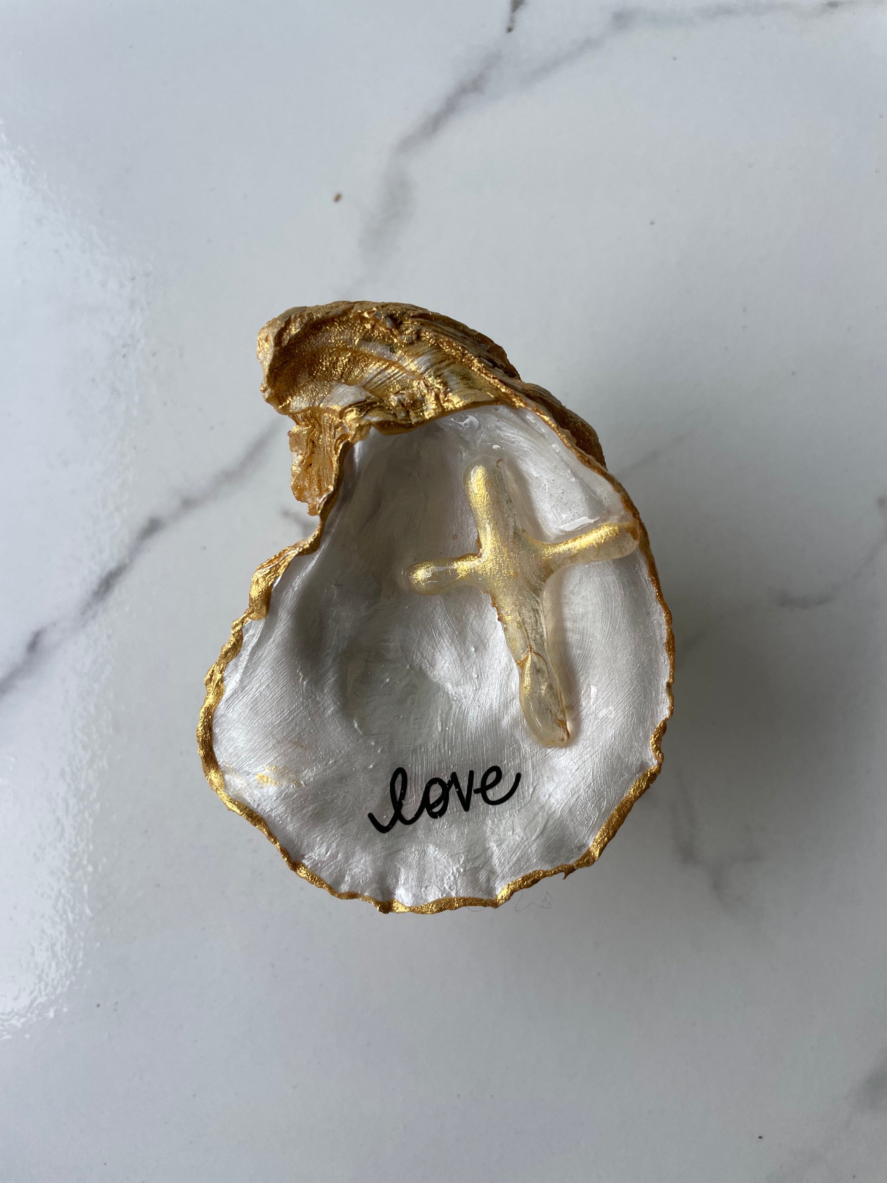 Love" Inspiration Series gilded oyster shell trinket dish on gold stand — hand-applied typographic lettering with dimensional gold cross accent, handmade by The Golden Shoreline
