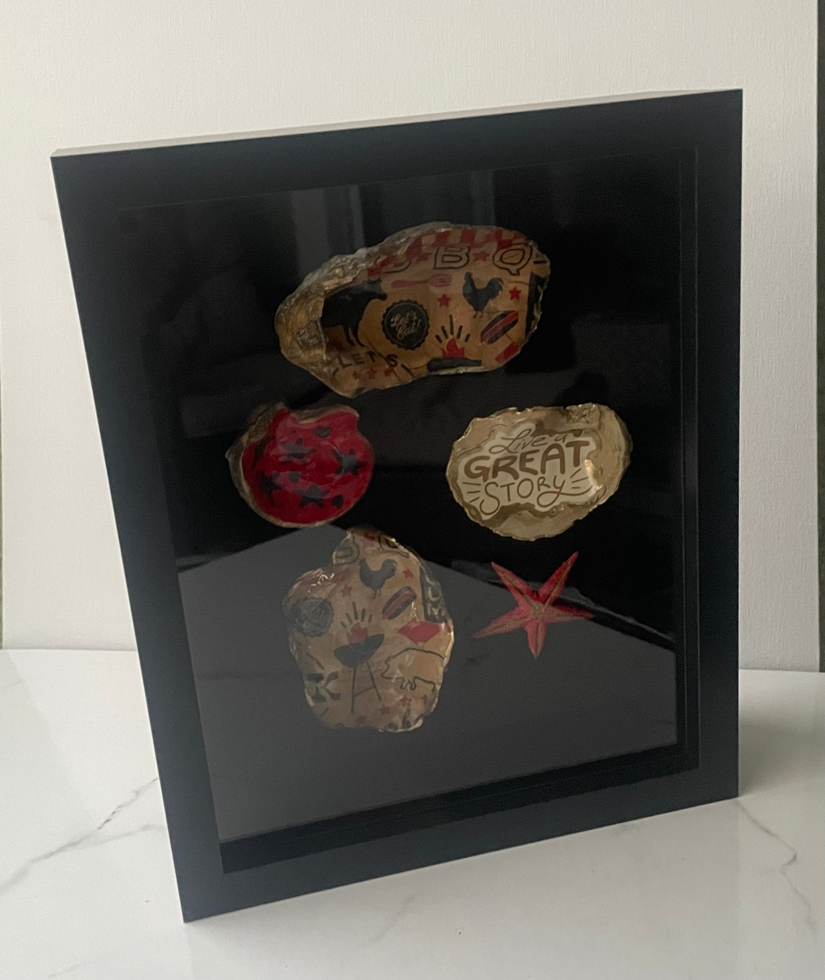 Close-up photo of the Shoreline Anthology gilded oyster shells showing BBQ motif, red star pattern, folk-art, and Live a Great Story designs