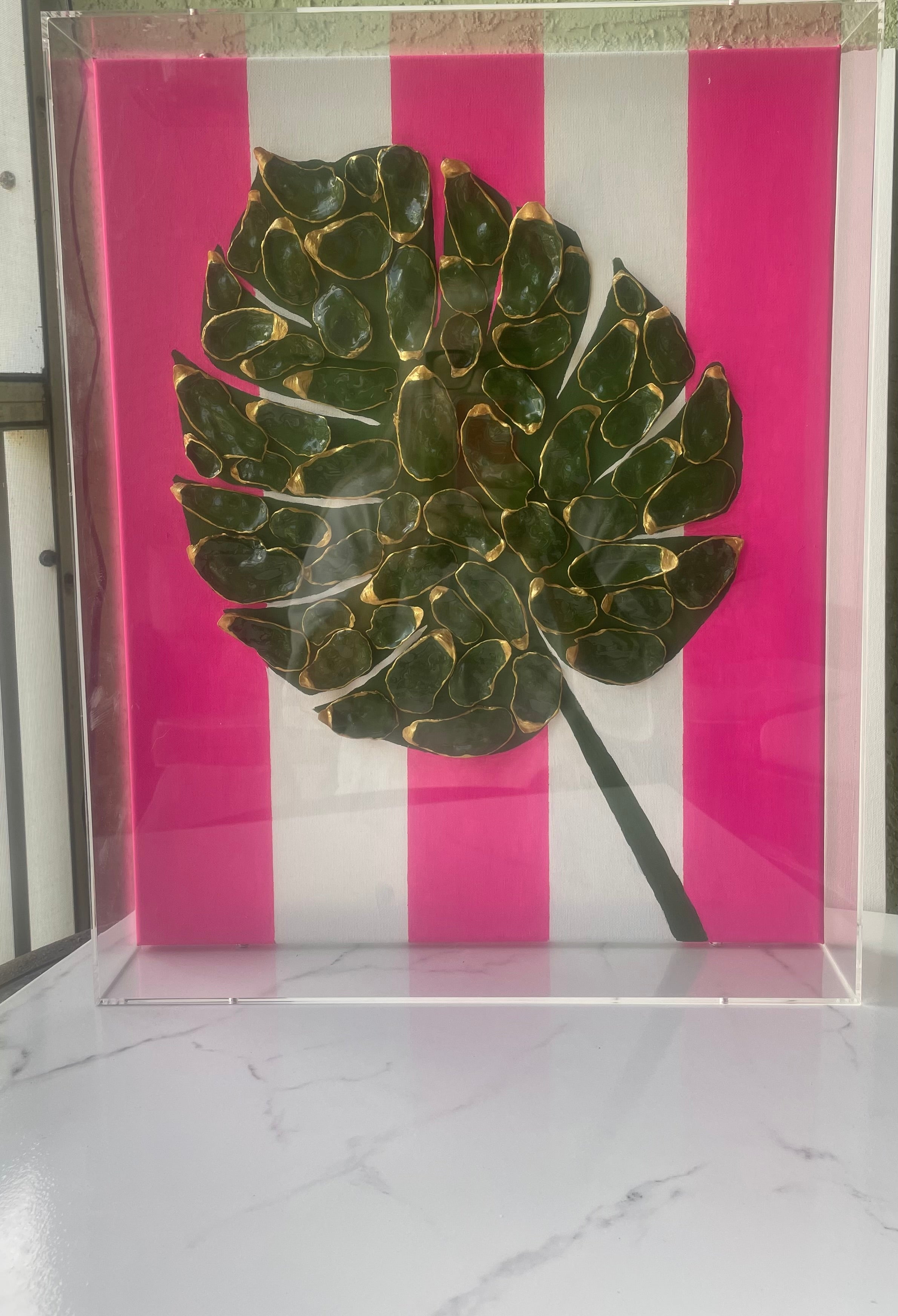 Gilded Monstera gallery wall art displayed outdoors — sculptural monstera leaf made from gold paint-edged shells on pink and white striped canvas in acrylic frame by The Golden Shoreline